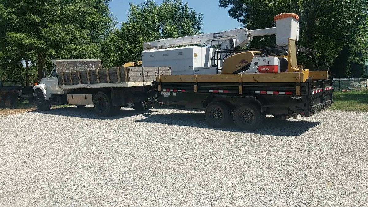 A bucket truck with a dump trailer carrying a compact loader, parked on gravel.